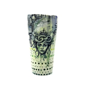 May include: A tall, cylindrical ceramic cup with a Medusa illustration. The cup features a black and white drawing of Medusa's face with a crown, set against a background of geometric shapes. The bottom half has green and black polka dots.
