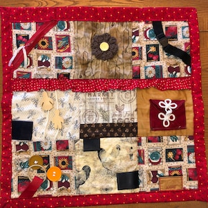 May include: A multi-textured sensory quilt with various fabrics, including a red zipper, a brown flower, a black buckle, a red pocket with white loops, and a brown fabric with the text "MARKET OPEN DAILY" and "Fury Fresh EGGS".