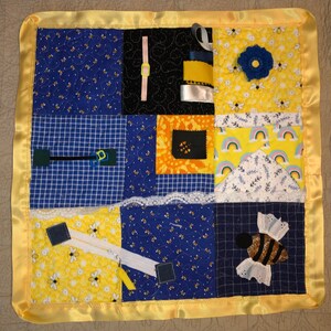 Könnte beinhalten: Eine gelbe und blaue Baby-Aktivitätsdecke mit verschiedenen Texturen und Merkmalen, darunter ein Reißverschluss, eine Blume, eine Biene und Bänder. Die Decke ist mit einem gelben Satinband eingefasst.