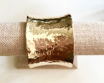 Puede incluir: Un brazalete dorado con textura martillada. El brazalete tiene forma curva y se muestra sobre un fondo de tela beige. El brazalete es una pieza de joyería ancha y llamativa.