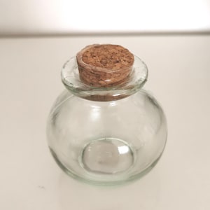 May include: A clear glass bottle with a round body and a cork stopper. The bottle has a wide, clear glass rim. The cork is a light brown colour and is textured. The bottle is on a white surface.