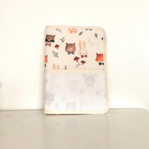 May include: A cream-coloured baby changing mat with a whimsical woodland animal print. The print features foxes, bears, and rabbits wearing bow ties, along with mushrooms and leaves. The mat has a white mesh pocket and a cream border.