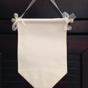 Pode incluir: Um banner de lona branco em branco com pinos de madeira e fita branca pendurado em um gancho de metal preto. O banner tem uma forma retangular com uma parte inferior pontiaguda.