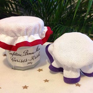 Può includere: Due coperchi per vasetti in tessuto bianco con bordi rossi e viola. Il coperchio del vasetto a sinistra ha un bordo rosso e dice "Confiture Bonne Cerises-Kirsch 3704".