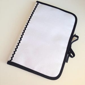 Peut inclure: Pochette blanche zippée avec une bordure noire et un ruban noir. La pochette est en tissu texturé.