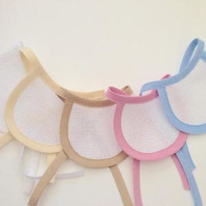 Peut inclure: Quatre bavoirs pour bébé faits à la main avec un tissu en maille blanche et une bordure de couleur. Les bavoirs sont beige, rose et bleu.