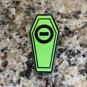 May include: A green coffin-shaped patch with a black circle and a horizontal line inside. The patch has black stitching around the edges.