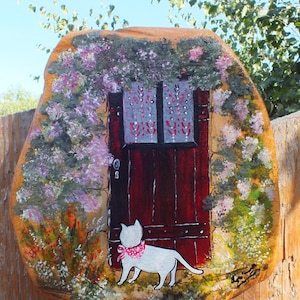 Peut inclure: Peinture sur pierre représentant un chat blanc avec un collier rose debout devant une porte rouge avec une fenêtre. La porte est recouverte d'une vigne avec des fleurs violettes.