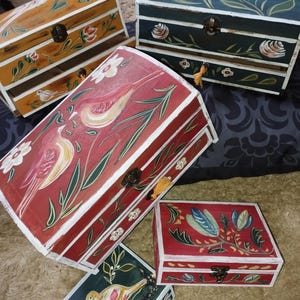 Può includere: Diverse scatole di legno dipinte a mano con motivi floreali e di uccelli. Le scatole sono di varie dimensioni e colori, tra cui rosso, verde e giallo, con bordi bianchi e chiusure in metallo. Le scatole hanno un'estetica vintage e artigianale.