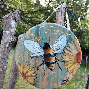 Op de afbeelding: Ronde houten plank met een handgeschilderde bij en zonnebloemen. De bij heeft zwart-gele strepen met witte vleugels. Het bord heeft een turquoise en oranje achtergrond met gouden accenten en een touwhanger.