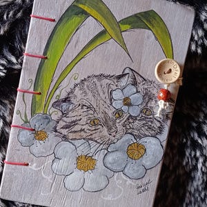 Op de afbeelding: Een wit houten dagboek met een handgeschilderde illustratie van een kat met bloemen. Het dagboek heeft rode stiksels en een houten knoop met de tekst "fait main" erop.