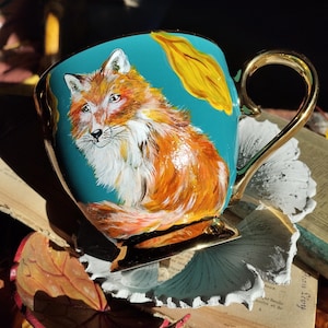 Peut inclure: Tasse à thé peinte à la main avec une illustration de renard. La tasse a un fond turquoise, une anse et des bordures dorées. Le renard est représenté en orange, blanc et marron. Une feuille jaune est peinte sur le côté de la tasse.