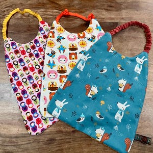 Könnte beinhalten: Drei bunte Baby-Lätzchen mit verschiedenen Mustern. Das erste Lätzchen ist rosa mit einem sich wiederholenden Muster von russischen Matroschkas. Das zweite Lätzchen ist weiß mit einem sich wiederholenden Muster von Tieren und Vögeln. Das dritte Lätzchen ist blau mit einem sich wiederholenden Muster von Eichhörnchen und Vögeln.