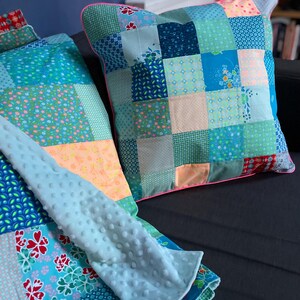 Peut inclure: Une courtepointe en patchwork avec un motif bleu, vert et rose. La courtepointe est composée de carrés de tissus différents, chacun avec un motif unique. La courtepointe est pliée en deux et placée sur une surface noire. Il y a un oreiller assorti avec une bordure rose.