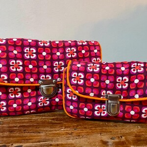 Könnte beinhalten: Zwei kleine Handtaschen mit einem geometrischen Blumenmuster in Rosa-, Rot- und Weißtönen auf dunkelviolettem Hintergrund. Die Handtaschen haben einen orangefarbenen Besatz und silberne Verschlüsse.