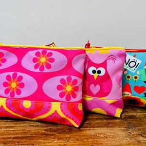 Peut inclure: Trois pochettes rectangulaires à fermeture éclair. La première est rose avec des motifs de fleurs orange et jaunes et une breloque violette. La deuxième est rose avec un hibou stylisé. La troisième est bleu clair avec des nains de jardin.