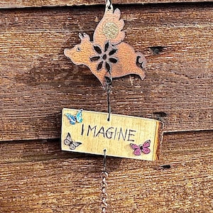 Peut inclure: Une décoration suspendue composée de trois perles en bois géométriques, d'une silhouette de cochon volant couleur cuivre avec une fleur, et d'une plaque en bois rectangulaire portant le mot IMAGINE et trois papillons, terminée par une clochette.