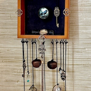 May include: A square wooden wall hanging featuring a black interior display with a circular bird image and a gold key. Below, multiple black cords suspend various charms including metal bells, a crescent moon, a clock face, and a gold cone bell.