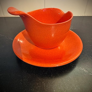 May include: An orange, speckled gravy boat with a matching saucer. The gravy boat has a curved handle and a wide, open bowl. The saucer is round and flat. Both pieces are made of a glossy material, likely ceramic or plastic.