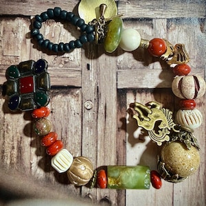 May include: A multi-strand bracelet featuring a mix of materials including matte black beads, a brass leaf charm, a large square multi-coloured stone centrepiece, red and cream-coloured beads, and a gold-toned metal horse-shaped charm.