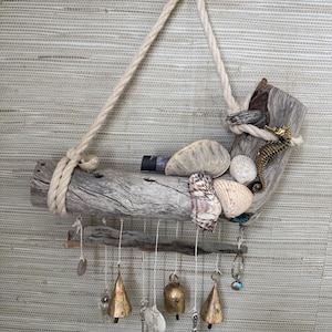 May include: A wall hanging made of two pieces of driftwood connected by white rope. The top piece is decorated with various seashells and a gold-colored seahorse figure. Hanging from the bottom piece are several metal bells, charms, and a small mirror.