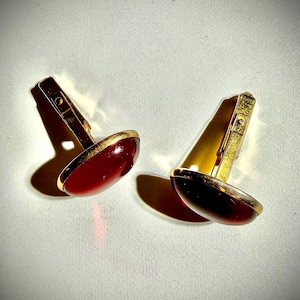 May include: A pair of gold-toned cufflinks with a deep red, oval-shaped center. The cufflinks have a gold border and a hinged clasp. The background is a light, neutral color.