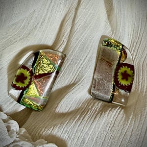 Puede incluir: Dos colgantes de vidrio dicroico con forma curva y transparente. Cada colgante presenta una mezcla de colores dorados, verdes y rojos, con un diseño floral. Los colgantes se exhiben sobre una tela texturizada de color blanquecino.