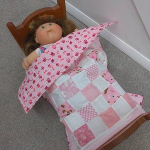May include: A pink and white patchwork quilt with a pink and white paw print blanket on top. A doll with green eyes and brown hair is lying in the bed.