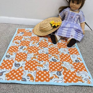 May include: A small, orange and blue quilted blanket with a fox print. The blanket is on a light brown carpet. A doll wearing a light blue dress and a straw hat is sitting on the blanket.