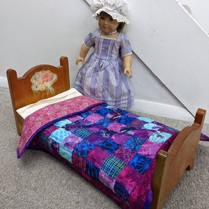 May include: A wooden doll bed with a floral painted headboard and a purple and blue patchwork quilt. The bed is made for a doll and is sitting on a gray carpet.