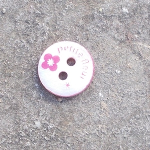 Può includere: Bottone bianco con un fiore rosa e il testo "Petite Neu" in rosa.