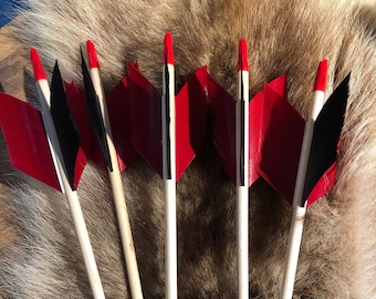 May include: Six wooden arrows with red and black fletching. The arrows are arranged in a fan shape and are lying on a brown fur surface.