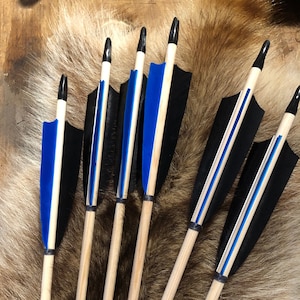 Op de afbeelding: Zes houten pijlen met zwarte en blauwe veren. De pijlen hebben een natuurlijke houten afwerking en zijn met een blauw lint aan elkaar gebonden.