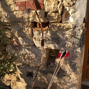 Op de afbeelding: Een houten boog met zwarte en bruine accenten en een zwarte snaar. De boog leunt tegen een stenen muur. Twee witte pijlen met rode veren leunen tegen de muur naast de boog.