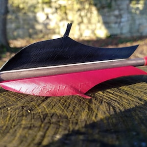 Op de afbeelding: Een close-up van een pijl met zwarte en rode veren. De pijl ligt op een houten oppervlak.