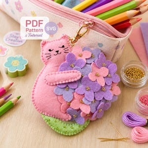 May include: A pink felt cat-shaped keychain holding a bouquet of purple and pink felt flowers. The keychain is attached to a pencil case filled with colored pencils. Other craft supplies include erasers, stickers, and embroidery floss.