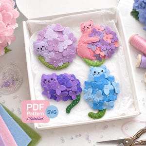 May include: A collection of handmade felt cat brooches, each nestled within a cluster of felt flowers. The brooches are in shades of pink, purple, and blue, with green stems. The image includes spools of thread, scissors, and felt fabric.