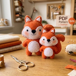 May include: Two orange felt fox plush toys, one large and one small, are displayed. The foxes have white bellies, faces, and ear tips, with black eyes and noses. Gold scissors and thread spools are on the wooden surface.