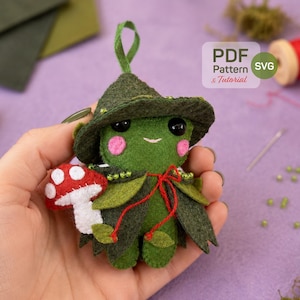 May include: A handmade felt frog doll wearing a witch hat and cape, holding a mushroom. The frog is green with pink cheeks and black eyes. The mushroom is red with white spots. The doll has a red bow and green leaves.