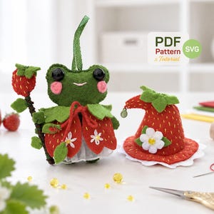 May include: A felt frog figurine wearing a strawberry-themed outfit and holding a strawberry staff, accompanied by a matching strawberry-shaped hat. The image includes a label that reads PDF Pattern & Tutorial SVG.