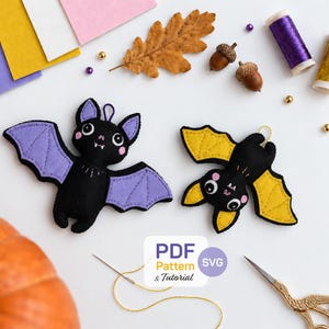 May include: Two felt bat ornaments, one with purple wings and one with yellow wings, featuring white fangs and pink cheeks. The image includes a PDF Pattern & Tutorial label, felt sheets, acorns, spools of thread, a pumpkin, and gold scissors.