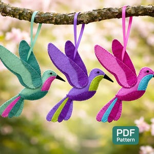 May include: Three felt hummingbird ornaments in shades of green, purple, and pink hang from a tree branch. Each bird has a contrasting belly and tail feathers, with black beaks and ribbon hangers. The background is a soft blur of pink and green.