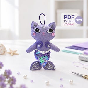 May include: A purple felt cat mermaid ornament featuring a sequined tail in shades of blue and purple, a white pearl waist accent, black bead eyes, and a small hanging loop. The image includes a label that reads PDF Pattern & Tutorial.