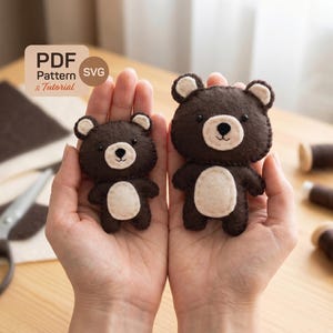 May include: Two handmade brown felt bear ornaments with white accents, held in open hands. The bears have white bellies and ear details. The image includes sewing supplies and text that reads "PDF Pattern & Tutorial SVG".