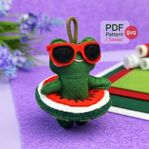 May include: A green felt frog ornament wearing red-framed sunglasses with black lenses. The frog is inside a watermelon-themed floatie with red, white, and green colors. A small brown loop is attached to the top. Text in the image reads PDF Pattern & Tutorial.
