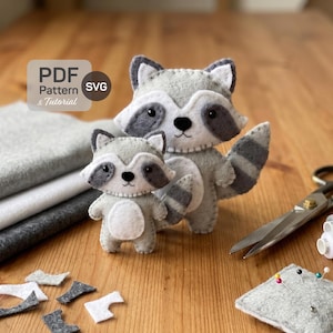May include: Two gray felt raccoon plushies with black eyes and striped tails. The image includes gray and white felt fabric, scissors, spools of thread, and a pin cushion. Text in the upper left corner reads "PDF Pattern & Tutorial SVG".