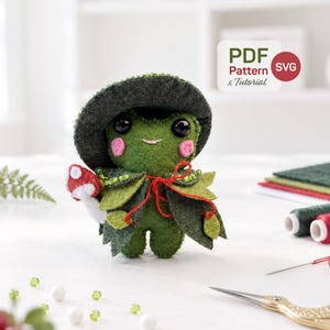 Puede incluir: Una muñeca de rana de fieltro verde con un sombrero verde oscuro y una capa en forma de hoja con lazos rojos. La rana tiene ojos negros, mejillas rosadas y sostiene un pequeño hongo rojo y blanco. Una etiqueta dice PDF Pattern & Tutorial y SVG.
