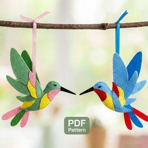 May include: Two felt hummingbird ornaments hanging from a branch. One is green, pink, and yellow, the other is blue, red, and yellow. Both have black beaks and are suspended by ribbons.