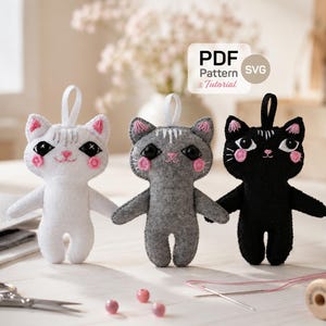 May include: Three felt cat ornaments in white, grey, and black, each featuring embroidered facial details, pink cheeks, and a hanging loop. A white label displays the text PDF Pattern & Tutorial SVG. Sewing tools and beads are visible nearby.