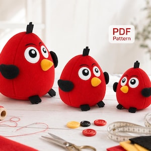 May include: Three plush red bird toys with black wings and feet, yellow beaks, and white eyes. The toys are in different sizes. The image also shows sewing supplies, including thread, buttons, and a measuring tape. A sign says "PDF Pattern".